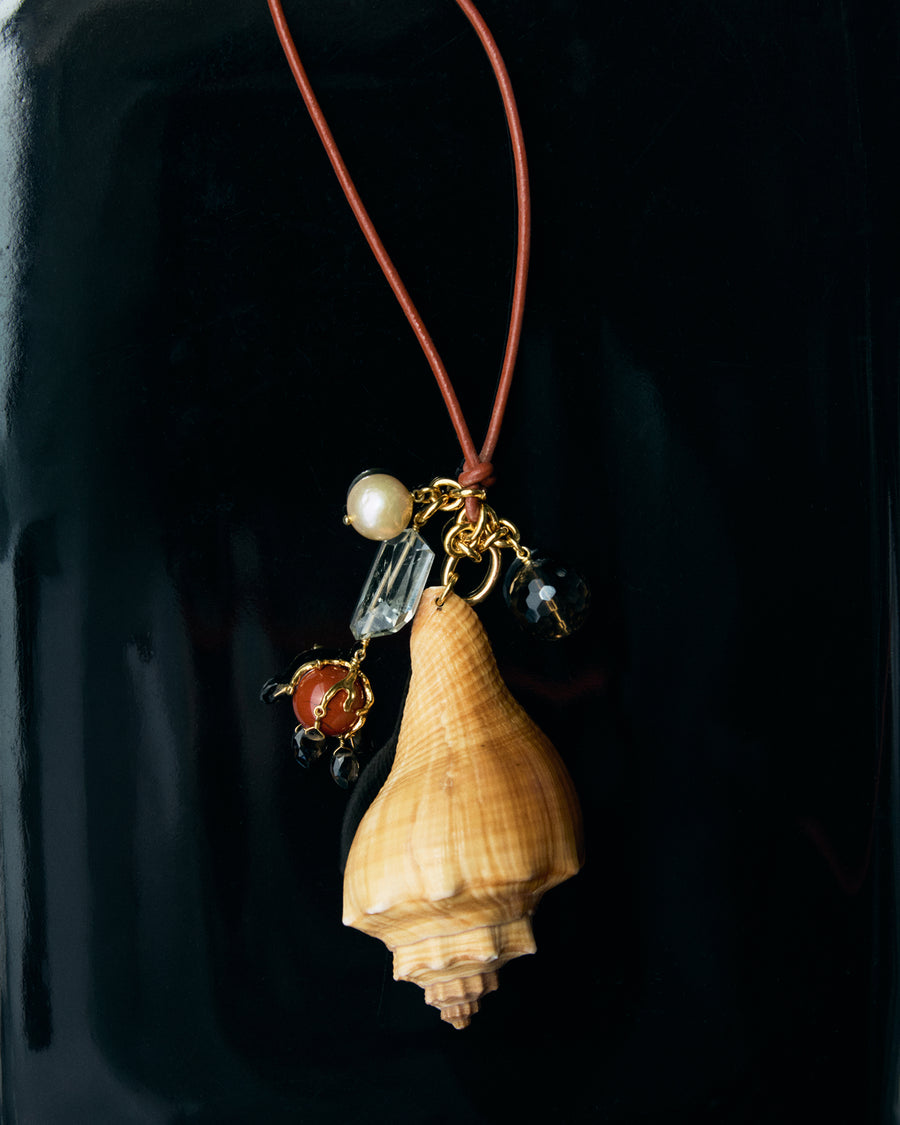 A handmade pendant necklace featuring a natural beige conch shell, gold-toned metal, a white pearl bead, a clear faceted crystal, and a small red-orange bead, suspended from a reddish-brown cord against a dark background.