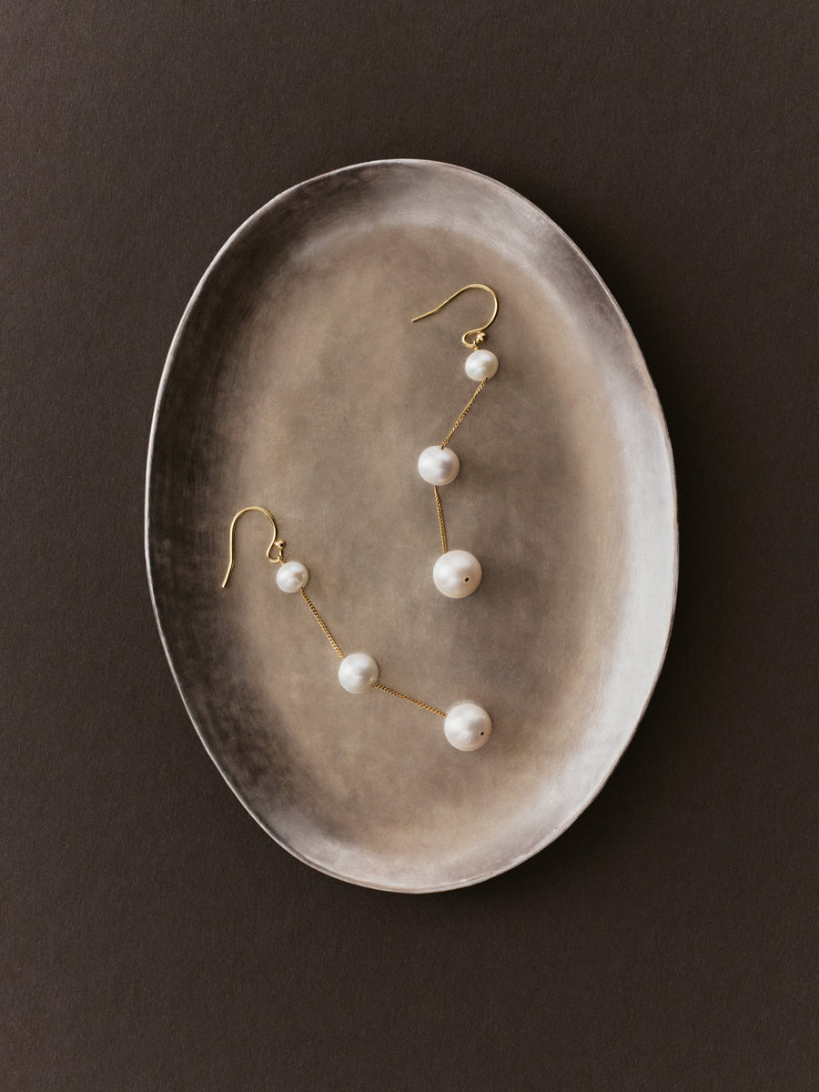 Gold drop earrings with white pearl beads, displayed on a textured oval ceramic dish against a dark brown background.