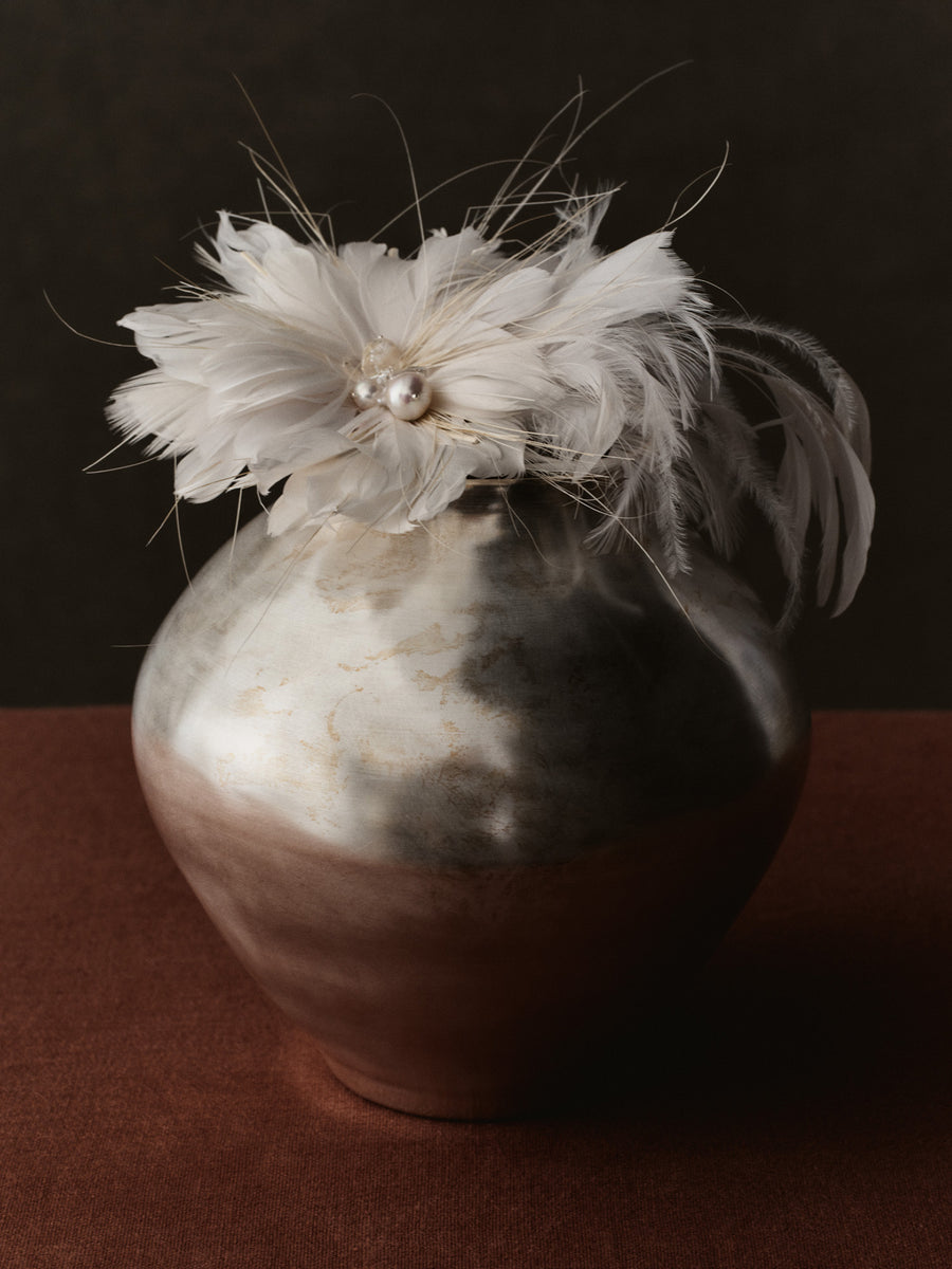 A decorative ceramic vase with a glossy metallic silver upper half and a warm brown lower half, topped with a large white feathered flower brooch with a small pearl accent, set against a dark brown background.